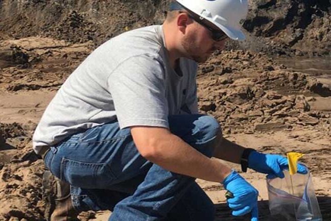 Technician in a hard hat and gloves taking a soil sample on a job site
