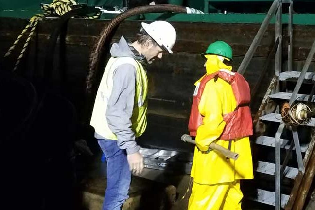 Acute Safety rep in a hard hat and vest supervising a worker in rain gear and life vest on a barge